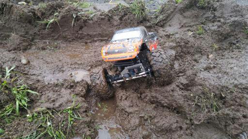 A radio-control monster truck driving through the deep mud. The truck is black, orange, and red with big tires. It's wet out. 