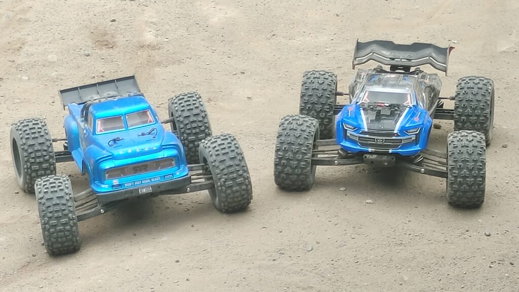 A blue ARRMA Kraton and blue ARRMA Notorious sitting side by side on a dirt RC car track.